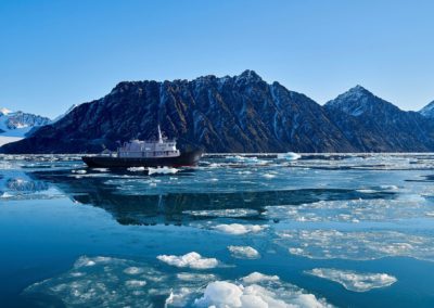 MS Balto navigiert durchs Treibeis mit teilweise schneebedeckten Bergen im Hintergrund