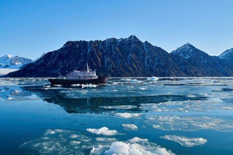 MS Balto navigiert durchs Treibeis mit teilweise schneebedeckten Bergen im Hintergrund